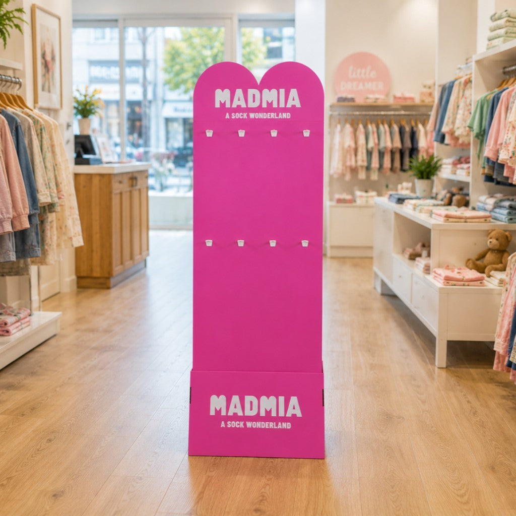 A bright pink cardboard display stand with the brand name 'MADMIA' and the text 'A SOCK WONDERLAND' printed in white, placed on a wooden floor inside a clothing boutique.